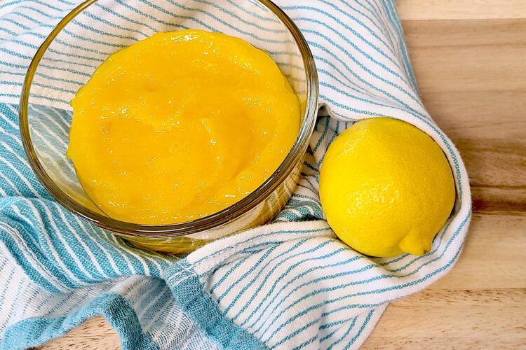 lemon curd in a glass bowl on a teal striped towel with a lemon on a wooden cutting board