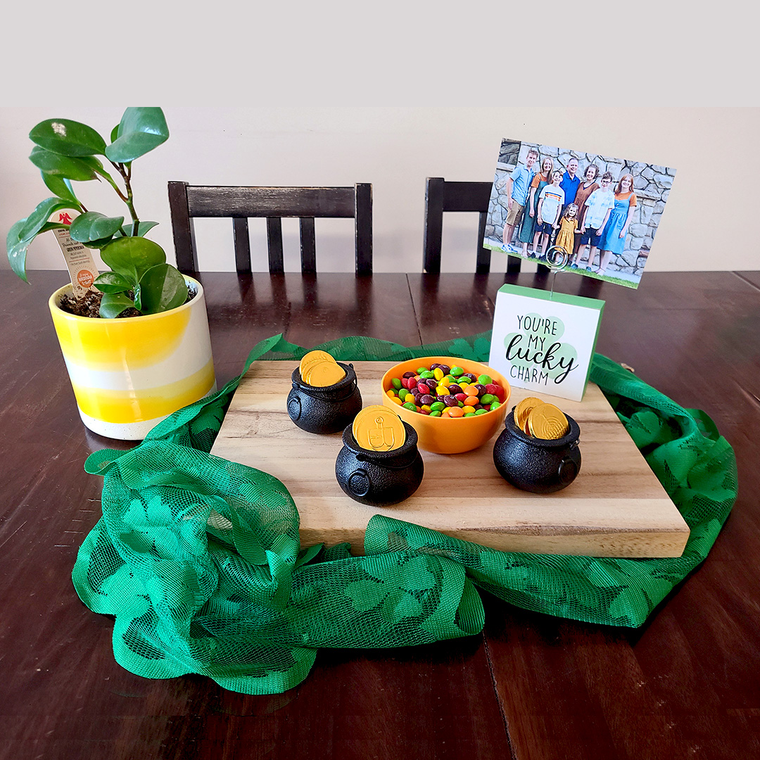 Skittles in an orange bowl along with gold chocolate coins in little black pots on a wooden board with sharmrock fabric under that