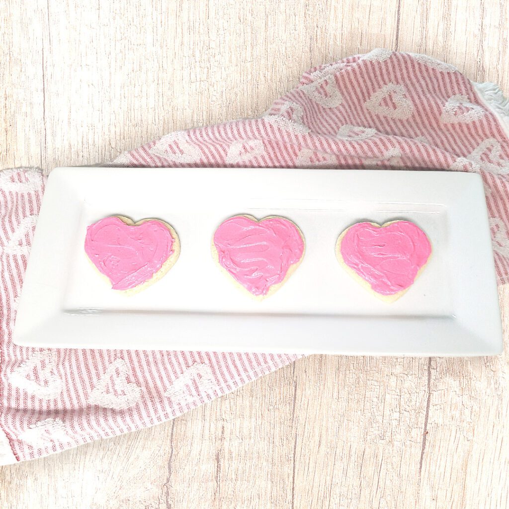 heart shaped sugar cookies with pink frosting on a white rectangular plate with a hand towel under the plate