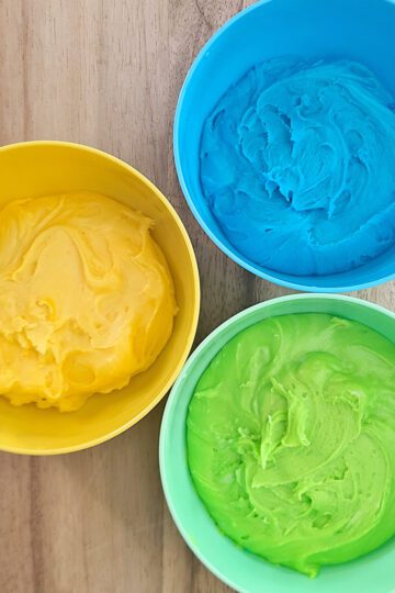 yellow, blue, and green buttercream frosting in coordinating colored bowls