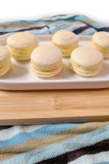 six lemon macaroons on a white oval dish on a wood cutting board with a striped towel underneath