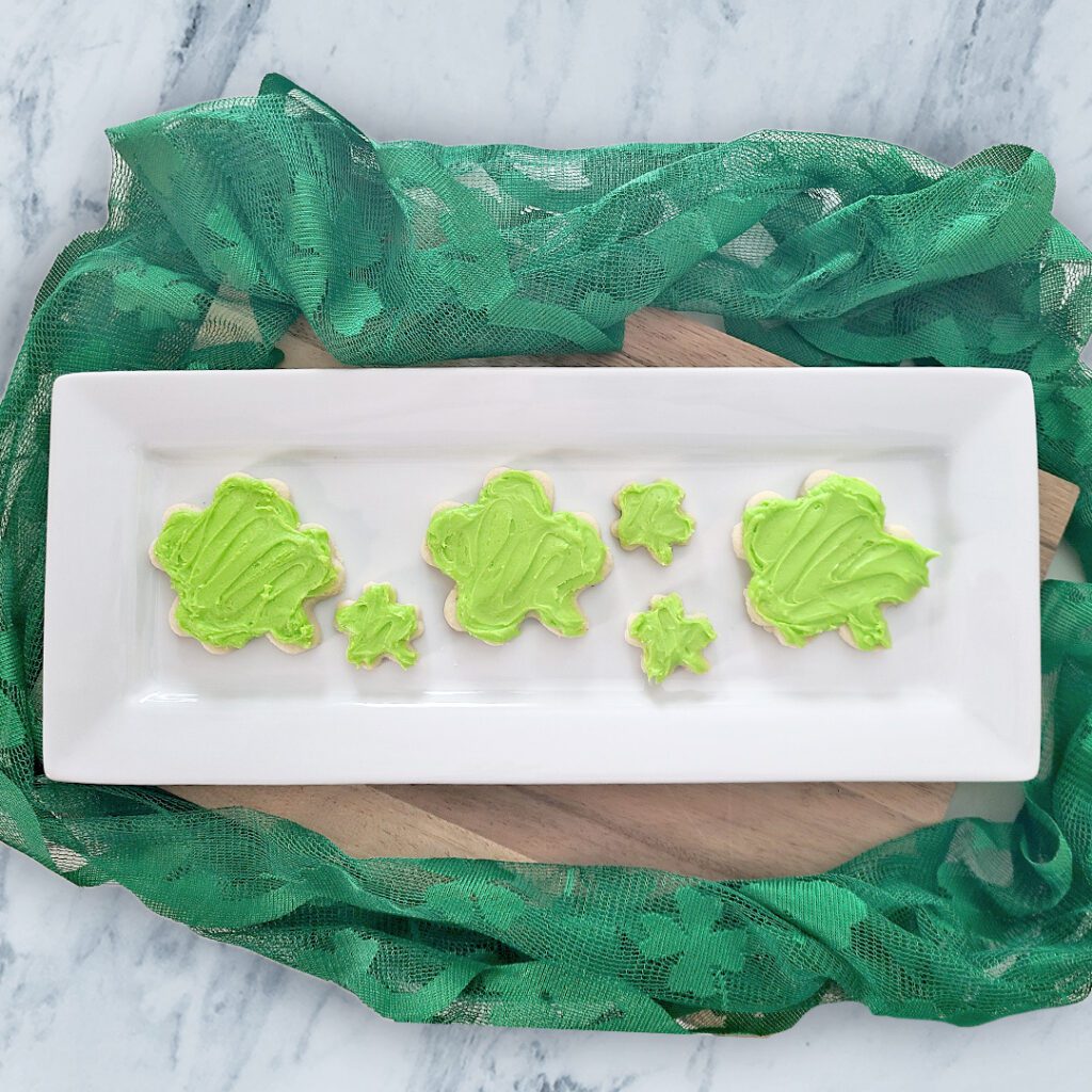 shamrock shaped gluten free sugar cookies with lime green frosting on a white rectangluar platter with green lace around it