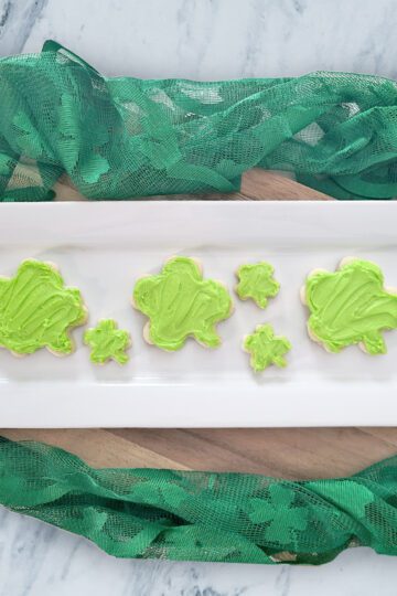 shamrock shaped gluten free sugar cookies with lime green frosting on a white rectangluar platter with green lace around it