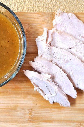 Herb roasted turkey slices next to a bowl of homemade gravy on a wooden cutting board