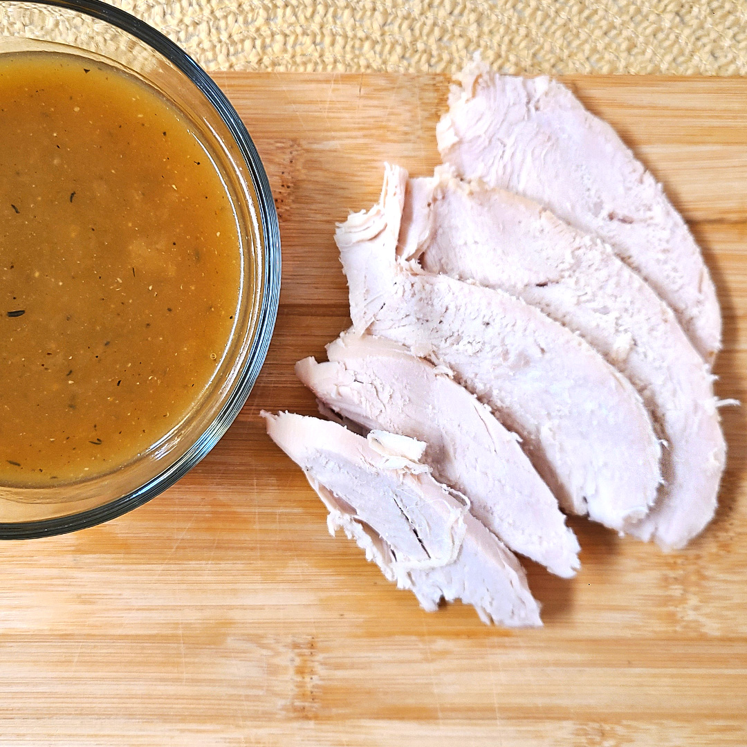 Herb roasted turkey slices next to a bowl of homemade gravy on a wooden cutting board