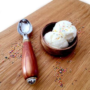 Homemade vanilla ice cream in a wooden bowl next to a ice cream scoop with sprinkles around on a cutting board