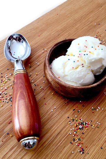 Homemade vanilla ice cream in a wooden bowl next to a ice cream scoop with sprinkles around on a cutting board
