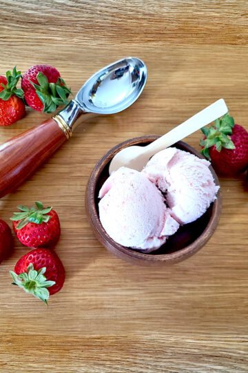 strawberry ice cream in a wooden bowl surrounded by fresh strawberries and a antique ice cream scoop all on a wooden board