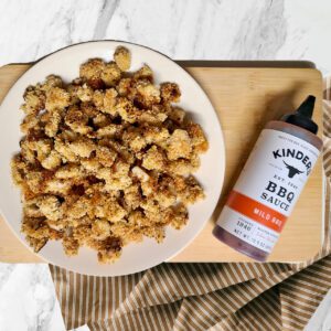 popcorn chicken on a white plate on top of a wooden cutting board with a bottle of Kinders BBQ sauce