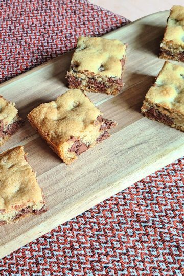 Gluten free chocolate chip cookie bars sitting on an oval wooden cutting board with a red table runner underneath