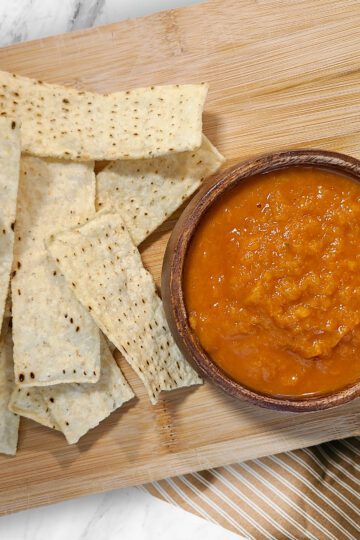 a small bowl of fresh peach & pepper salsa on a wooden cutting board with tortilla chips around it