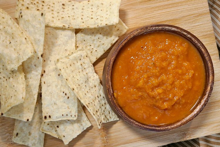 a small bowl of fresh peach & pepper salsa on a wooden cutting board with tortilla chips around it