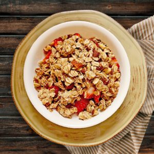 Cinnamon Vanilla granola in a white bowl with fresh strawberries on top of a round light brown plate