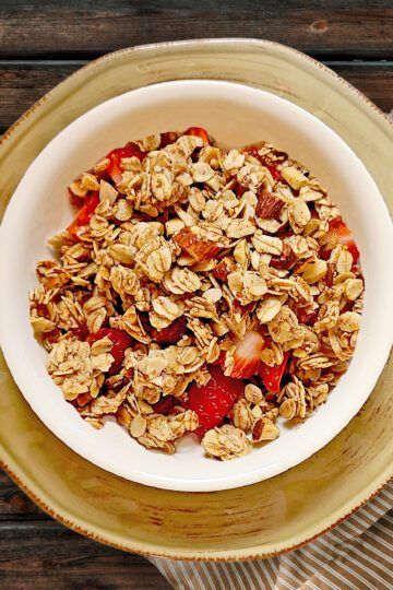 Cinnamon Vanilla granola in a white bowl with fresh strawberries on top of a round light brown plate