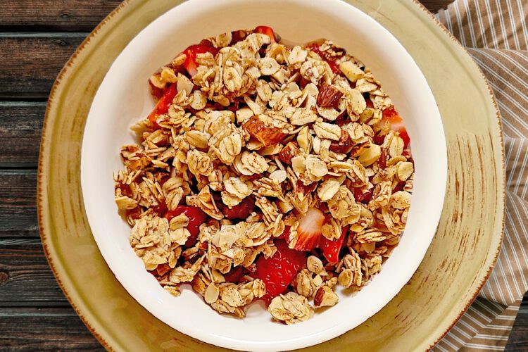 Cinnamon Vanilla granola in a white bowl with fresh strawberries on top of a round light brown plate